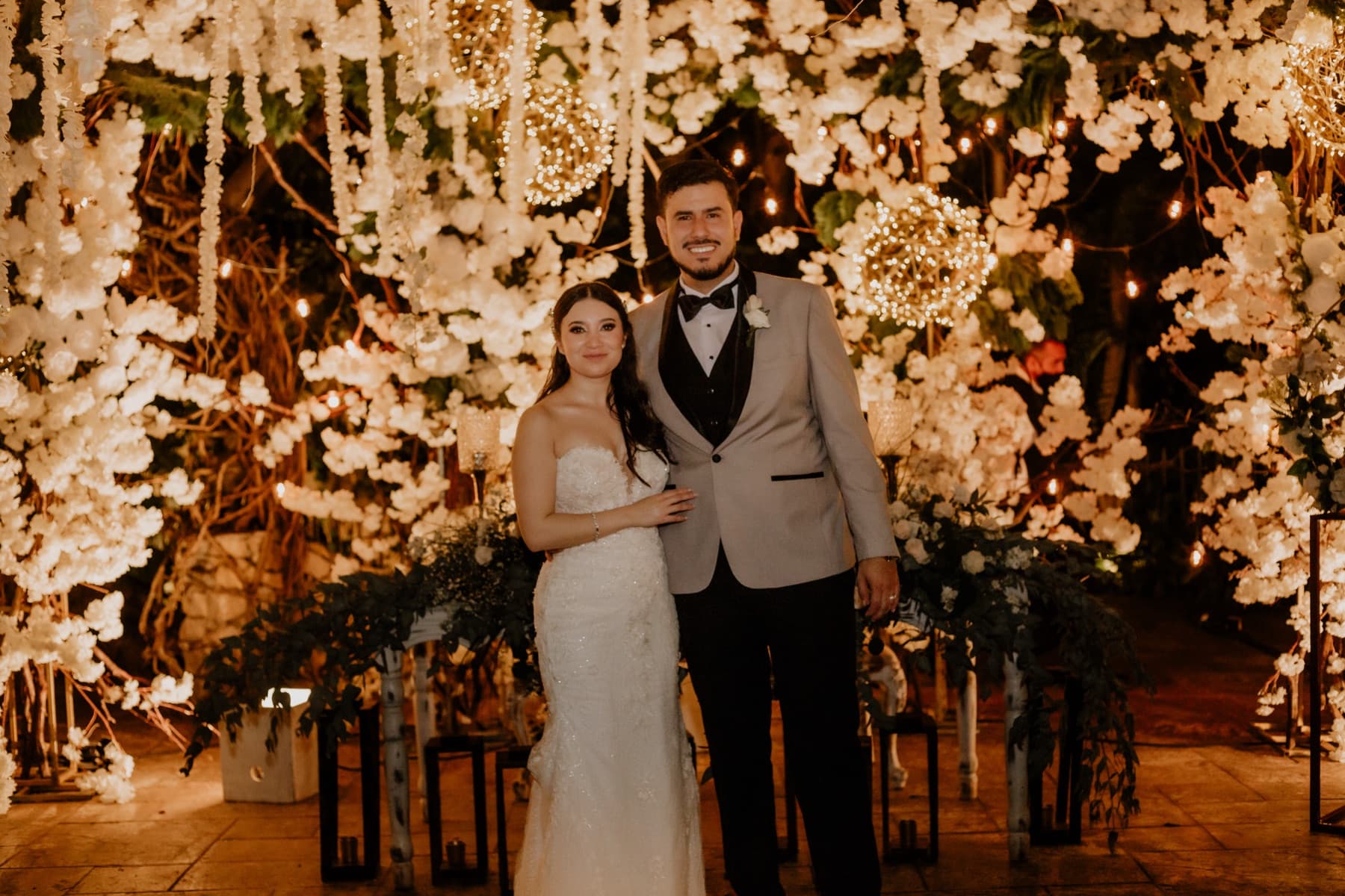 Andrea y Andrés el día de su boda, frente a un muro de flores blancas iluminado.